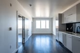 This single-floor space presents a clean, contemporary design characterized by bright white walls, smooth ceilings, and rich dark hardwood flooring that runs throughout the main living areas and bedrooms. The open-plan layout connects the living area directly to the kitchen, creating a cohesive and functional space that feels airy and well-proportioned.

The kitchen is outfitted with flat-front cabinetry in a soft grey finish, a dark glass tile backsplash, light stone countertops, a stainless steel sink, a gas cooktop, and a built-in oven — offering a sleek, modern aesthetic with practical utility. Ample upper and lower cabinet storage is provided along the full length of the kitchen wall.

The living area benefits from generous natural light streaming through large windows, which frame elevated views of the surrounding neighborhood, rooftops, mature trees, and rolling hills under open skies. Additional rooms share this same bright, minimalist character, each featuring the same dark hardwood floors and large windows with outdoor views.

The bathroom maintains the contemporary theme with dark mosaic tile flooring, a floating white vanity with a wide mirror, and a full tub and shower combination clad in white subway tiles. Throughout, the space delivers a cohesive, modern aesthetic with quality finishes.