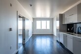This single-floor space presents a clean, contemporary design characterized by bright white walls, smooth ceilings, and rich dark hardwood flooring that runs throughout the main living areas and bedrooms. The open-plan layout connects the living area directly to the kitchen, creating a cohesive and functional space that feels airy and well-proportioned.

The kitchen is outfitted with flat-front cabinetry in a soft grey finish, a dark glass tile backsplash, light stone countertops, a stainless steel sink, a gas cooktop, and a built-in oven — offering a sleek, modern aesthetic with practical utility. Ample upper and lower cabinet storage is provided along the full length of the kitchen wall.

The living area benefits from generous natural light streaming through large windows, which frame elevated views of the surrounding neighborhood, rooftops, mature trees, and rolling hills under open skies. Additional rooms share this same bright, minimalist character, each featuring the same dark hardwood floors and large windows with outdoor views.

The bathroom maintains the contemporary theme with dark mosaic tile flooring, a floating white vanity with a wide mirror, and a full tub and shower combination clad in white subway tiles. Throughout, the space delivers a cohesive, modern aesthetic with quality finishes.