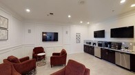 This single-floor coffee bar is thoughtfully designed to blend functionality with a comfortable lounge atmosphere. The space is divided into two distinct zones: a relaxed seating area and a well-equipped beverage station. The seating area features plush, deep-red armchairs arranged around a decorative side table, creating an inviting setting for casual conversation or quiet downtime. A large wall-mounted flat-screen TV is centered on a white paneled accent wall, adding a refined architectural detail that elevates the room's aesthetic.

Adjacent to the lounge area, the beverage station is outfitted with dark cabinetry, a granite tile backsplash, and a lineup of appliances including a coffee maker, microwave, and a compact refrigerator. A second wall-mounted TV is positioned above the counter for added convenience. Recessed lighting throughout the space provides bright, even illumination, complemented by light-toned tile flooring that enhances the clean, modern feel. Crown molding along the ceiling adds a polished finishing touch, making this coffee bar an appealing and well-appointed communal space.