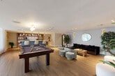 Located at 1000 Sunnybrook Rd in Miami, FL, this thoughtfully designed single-floor space offers a sophisticated blend of leisure and functionality. The open layout is anchored by warm wood-look flooring that flows seamlessly throughout, creating a cohesive and inviting atmosphere.

The living area is generously appointed with a plush dark velvet sectional sofa, curved upholstered accent chairs, and ottomans arranged around a wood coffee table. A large flat-screen TV is mounted on a crisp white wall, while a patterned accent wall adds texture and visual depth. An oval mirror, gold wall sconce, and tall indoor plants lend warmth and a touch of organic elegance. A full-size pool table serves as a central entertainment feature, making the space ideal for social gatherings.

The kitchen area is refined and well-equipped, showcasing a striking island with a white stone countertop and ribbed blue cabinetry with brushed gold hardware. Built-in open shelving in warm wood tones displays curated décor and a coffee station. Pendant lighting and recessed ceiling lights provide a layered, welcoming glow.

Large glass double doors invite natural light and connect the interior to an outdoor area, enhancing the overall sense of openness. Contemporary artwork throughout adds a curated, gallery-like character to this polished Miami retreat.