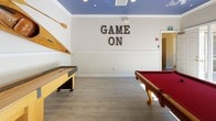 This well-appointed game room delivers a lively recreational environment designed for socializing and entertainment. The space is anchored by a classic red-felt pool table and a full-size shuffleboard table, providing ample opportunity for friendly competition. A striking blue ceiling with recessed lighting adds a bold design element, contrasting with the neutral wall tones and crown molding that frame the room with a polished finish.

A decorative accent wall featuring a geometric patterned wallcovering serves as a focal point, hosting a wall-mounted flat-screen television above a built-in wet bar area. The bar includes dark cabinetry, a dedicated beverage refrigerator, and counter space, making it ideal for hosting gatherings. A wall display of decorative wooden kayaks and oars adds a nautical theme and visual interest throughout the space.

Wood-look flooring runs through much of the room, complemented by tile near the exterior entry. Large glass-paned doors flood the room with natural light and open directly to an outdoor patio, where a bistro-height table and chairs offer additional seating. A bold "GAME ON" wall sign reinforces the room's energetic and recreational character, creating a welcoming and spirited atmosphere.