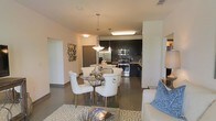 This single-story space showcases an open-concept layout that connects the living, dining, and kitchen areas into one cohesive and functional environment. The dining area is anchored by a round glass-top table surrounded by elegant white upholstered chairs with nail-head trim detailing, suspended beneath a pair of sleek pendant lights that add a refined focal point to the space. Decorative wall art adds a touch of personality to the adjacent wall.

The kitchen is well-appointed with dark cabinetry, stainless steel appliances including a range and built-in microwave, and a linear overhead fluorescent fixture that ensures ample task lighting. The dark lower cabinetry contrasts effectively with the lighter walls, creating a bold, modern aesthetic. A pass-through or peninsula-style counter connects the kitchen to the dining area, enhancing the open flow.

The living area, partially visible in the foreground, features a neutral-toned sofa accented with blue throw pillows, complemented by a patterned area rug. Wood-style flooring runs throughout, tying the spaces together with warmth and continuity. Overall, the space presents a modern, well-designed interior with cohesive finishes.