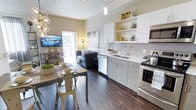 This thoughtfully designed single-floor residence blends contemporary style with warm, inviting details across its well-appointed living spaces. The bedroom is a standout feature, showcasing a tufted upholstered headboard, rich dark hardwood flooring, and a striking planked white ceiling with a ceiling fan that adds architectural interest overhead. Large windows allow natural light to pour in, offering views of the surrounding cityscape, while sheer curtains provide a soft, elegant touch. An industrial-style open shelving unit with chain-link accents serves as both a media stand and decorative display area, complementing the room's eclectic yet refined aesthetic. The open-concept kitchen, dining, and living area is equally impressive, featuring stainless steel appliances, light gray cabinetry, open upper shelving, and a classic subway tile backsplash. A single pendant light and a cascading globe chandelier add character and warmth to the space. The dining area accommodates a rustic wood table with vintage metal chairs, while the living area benefits from direct access to an outdoor balcony through sliding glass doors. Consistent dark hardwood flooring and planked white ceilings tie all areas together, creating a cohesive and polished interior throughout.