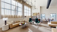 This thoughtfully designed combined kitchen and living space makes a striking first impression with its soaring ceilings and expansive floor-to-ceiling windows dressed in sheer, light-diffusing curtains that flood the interior with abundant natural light. A pair of sculptural, globe-adorned pendant chandeliers serve as dramatic focal points overhead, lending a contemporary and artistic character to the space.

The living area is elegantly appointed with a plush, neutral-toned sofa complemented by rich emerald green velvet swivel chairs, all arranged around sleek, tiered marble-top coffee tables atop a herringbone-patterned carpet. A wall-mounted flat-screen television is paired with a minimalist media console, while an additional seating nook near picture windows offers views of the surrounding exterior.

The kitchen area showcases warm walnut cabinetry accented with brass hardware, crisp white countertops, and stainless steel appliances. A bold three-dimensional geometric tile backsplash adds visual depth and artistry to the space. Linear LED lighting beneath upper cabinetry provides clean, modern illumination. A dedicated sink station with the same striking geometric tile backdrop and a water fountain add practical utility. A staircase with sleek railings connects to additional areas, reinforcing the multi-functional and communal nature of this well-appointed space.