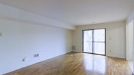 This two-floor space offers a clean, minimalist aesthetic defined by warm hardwood flooring and crisp white walls throughout. The main living area is generously proportioned, providing an open and airy feel that is enhanced by abundant natural light streaming in through a large sliding glass door. This door leads directly to an outdoor balcony, which overlooks a lush, tree-lined setting — a standout feature that brings the outdoors in and creates a serene atmosphere. The hardwood floors display a classic parquet-style pattern, adding warmth and character to the space. Subtle architectural details, such as a slightly recessed ceiling border and integrated HVAC vents, reflect a practical and well-considered design. The neutral white palette of the walls offers a versatile backdrop suited to a wide range of interior styles. Spanning two floors, the layout provides distinct zones for living and other functions, giving occupants both shared and private areas. Overall, the space balances simplicity and functionality, making it well-suited for those who value natural light, outdoor access, and timeless design elements.