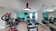 This fitness center offers a thoughtfully arranged workout space designed to accommodate a variety of exercise routines. The main workout area features wood-look flooring throughout, crown molding, and a soft gray color palette accented by a bold teal feature wall that adds energy to the environment. Multiple windows allow natural light to fill the space, complemented by ceiling fans and recessed lighting for comfortable year-round use.

The cardio section is equipped with several treadmills, a stationary bike, and a rowing machine, providing diverse options for aerobic training. A multi-station cable weight machine anchors the strength training area, while a full dumbbell rack with a range of free weights supports additional resistance workouts. A wall-mounted flat-screen television faces the cardio equipment, and a large wall mirror enhances the sense of space while allowing users to monitor their form.

A separate entry corridor connects to the main workout room via a French door and features dual water fountains for convenient hydration. The overall layout is clean and functional, offering a practical and comfortable environment suited for a well-rounded fitness routine.