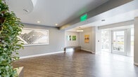 This thoughtfully designed lobby and common area presents a clean, contemporary aesthetic anchored by a dramatic cylindrical living green wall — a lush, floor-to-ceiling plant installation housed in a sleek metallic planter that serves as the visual centerpiece of the space. The surrounding area features warm wood-look flooring that extends throughout, complementing the soft gray and white wall tones that lend the space a calm, professional atmosphere.

Recessed and pendant lighting illuminate the open layout, which flows seamlessly between a main lobby zone, a transition corridor, and a welcoming lounge area furnished with upholstered seating and a low bench. Curated framed artwork lines the walls, adding visual interest and a sense of community identity. A circular ceiling detail frames the living wall from above, reinforcing its role as a focal point.

The space includes stainless steel elevator doors and an adjacent staircase with handrails, providing accessible multi-directional movement throughout. Glass entry doors at one end draw in natural light from outside, brightening the entry corridor. A dedicated seating alcove with a ring pendant light and oval wall art offers a comfortable waiting area. Overall, the space balances biophilic design elements with modern finishes to create an inviting, functional communal environment.