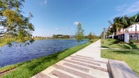 This single-level outdoor space offers a beautifully landscaped lakeside setting that combines natural beauty with thoughtful design. The centerpiece of the space is a generously wide decorative paver walkway, laid in a multi-toned brick pattern that adds visual interest and a polished, welcoming feel underfoot. The path runs alongside a large, calm lake, whose still waters reflect the surrounding greenery and open sky, creating a serene and picturesque atmosphere.

A central fountain rises gracefully from the lake, adding both visual appeal and the soothing sound of flowing water to the environment. The shoreline is framed by neatly maintained green grass and a variety of young and mature trees, including what appear to be palms and deciduous varieties, providing natural shade and seasonal color throughout the year. Decorative lamp posts line the walkway at intervals, suggesting the space is equally inviting during evening hours.

The open, flat layout allows for unobstructed views across the water toward a lush tree line and what appears to be a residential community in the background. The abundant natural light and expansive sky overhead contribute to an airy, open ambiance. This is a well-appointed outdoor environment ideal for leisurely walks and enjoying the natural surroundings.