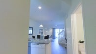 This two-story space is designed with an open-concept layout that integrates the kitchen, dining, and living areas into one cohesive, flowing environment. The kitchen is outfitted with granite countertops and a peninsula that serves as both a workspace and a casual dining surface, complemented by a white dishwasher and a sink. A pendant light hangs above the kitchen area, adding a focused decorative element to the space. The dining area sits adjacent to the kitchen and features seating for several guests, illuminated by an arc floor lamp that adds a modern touch. Beyond the dining area, the living room receives generous natural light through large windows or a glass door at the rear, creating a bright and welcoming ambiance throughout. Recessed ceiling lights and a ceiling fan provide additional illumination and comfort. The color palette throughout is light and neutral, with white walls and ceilings that enhance the sense of openness. A staircase visible toward the rear of the space connects to the second floor, reinforcing the home's multi-level character.
