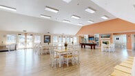 This single-story community clubhouse in Livermore, CA (94551) offers a bright, well-appointed gathering space designed to foster social interaction and recreation. The centerpiece is a expansive main hall featuring soaring vaulted ceilings and an array of skylights and flush-mounted fixtures that flood the space with abundant natural light. Light wood-tone flooring runs throughout, complementing the clean white walls and warm peach accent wall that adds a welcoming pop of color. Multiple dining tables with white cross-back chairs are arranged throughout the hall, accommodating group gatherings and events with ease. A dedicated recreation area houses a full-size billiards table, while a built-in shelving unit serves as a library nook stocked with books and topped with a wall-mounted TV and a decorative fireplace surround. A separate wing features additional seating and dining space alongside an accessible kitchen equipped with standard appliances, ideal for catered events or resident use. The entry foyer provides a comfortable lounge area with armchairs and artwork, creating a warm first impression. Large windows along the perimeter frame outdoor greenery and allow generous natural light throughout. Multiple exterior doors ensure easy access, making this clubhouse a functional and inviting hub for community life.