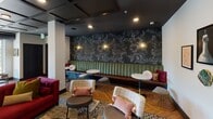 This single-floor space is thoughtfully designed as a multifunctional communal gathering area, seamlessly blending lounge, dining, and kitchen zones within one open-plan layout. Throughout, herringbone-patterned hardwood flooring provides a warm, cohesive foundation beneath the varied activity areas.

The lounge zone features a wall-mounted flat-screen TV positioned above a gold-toned media credenza, surrounded by curated wall art and an eclectic mix of seating including upholstered chairs, wire-frame accent stools, and patterned area rugs. A dramatic banquette seating section anchors another corner, lined in channel-tufted green velvet against a bold, dark floral wallcovering with antique-style brass sconces, creating an intimate atmosphere.

The kitchen area is highlighted by a large white waterfall island with tiled detailing, topped by globe pendant lights and framed by deep green paneled walls with a dark coffered ceiling overhead. Bar stools line the island, making it ideal for casual gathering.

Adjacent to the kitchen, a formal dining area accommodates a long wooden table beneath an ornate chandelier. Expansive floor-to-ceiling sliding glass doors flood the space with natural light and provide direct access to an outdoor terrace with additional seating.