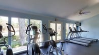 This single-level fitness center offers a dedicated space for cardiovascular exercise, outfitted with a selection of machines including ellipticals, an upright stationary bike, and treadmills. The room is bathed in natural light streaming through large windows that line the walls, providing pleasant outdoor views of surrounding greenery and trees. A set of glass French doors offers direct access to the exterior, further enhancing the open, airy feel of the space. The walls are painted in a soft blue-gray tone, complementing the light gray flooring and white crown molding that frames the ceiling. The layout is thoughtfully organized, with cardio equipment arranged to maximize floor space and allow comfortable movement between machines. A wall-mounted television is positioned for viewing during workouts, adding a practical amenity. The overall ambiance is clean, functional, and welcoming, with the abundance of natural light creating an energizing atmosphere suited for daily exercise routines.