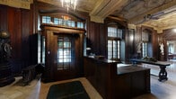 This distinguished lobby space exudes classical grandeur across its expansive single floor. The architectural design is immediately striking, defined by richly stained dark wood paneling that lines the walls from floor to ceiling, complemented by elaborately detailed coffered and gilded ceilings adorned with intricate plasterwork and geometric molding. Polished marble tile flooring runs throughout, reflecting the warm glow of crystal chandeliers and vintage pendant lanterns overhead.

The entry vestibule features a traditional revolving door framed in dark wood with multi-pane glass transoms, setting a formal tone from the moment of arrival. A staffed reception desk anchors the main hallway, which extends into a long, gallery-like corridor flanked by tall fluted columns. Classical marble and bronze sculptures are displayed on pedestals throughout, lending the space a museum-like quality.

The living room area offers a refined sitting space with ornate wall paneling in warm golden tones, decorative murals, and crystal chandelier lighting. Tufted leather seating and vintage upholstered armchairs provide comfortable furnishings beneath windows that allow natural light to filter in. A stately grandfather clock and gilded mirrors further reinforce the space's historic elegance. The overall ambiance is one of timeless, formal sophistication.