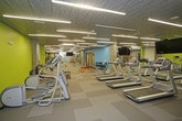 This contemporary fitness center is designed to accommodate a wide range of workout styles and fitness goals. The space is defined by its striking interior palette, featuring bold accent walls in lime green and aqua blue set against cool grey tones, creating an energizing atmosphere throughout. The textured dark ceiling panels paired with rows of linear LED strip lighting provide bright, even illumination across the entire floor area.

The facility is organized into distinct functional zones. A dedicated cardio area hosts multiple treadmills, stationary bikes, and rowing machines, all positioned near large wall-mounted mirrors and flat-screen TVs for an engaging workout experience. Adjacent to this is a strength training zone equipped with a multi-functional cable and power rack system, a punching bag, and a selection of barbells and dumbbells arranged on tiered racks.

The flooring throughout consists of durable, checkered grey carpet tiles that provide both comfort and traction. Exposed concrete columns add an industrial architectural character to the space. Additional amenities include a water fountain, ample open floor space for stretching or functional movement exercises, and a separate room accessible via a private door. Full-length mirrors along one wall visually expand the space and assist users during training sessions.