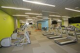 This contemporary fitness center is designed to accommodate a wide range of workout styles and fitness goals. The space is defined by its striking interior palette, featuring bold accent walls in lime green and aqua blue set against cool grey tones, creating an energizing atmosphere throughout. The textured dark ceiling panels paired with rows of linear LED strip lighting provide bright, even illumination across the entire floor area.

The facility is organized into distinct functional zones. A dedicated cardio area hosts multiple treadmills, stationary bikes, and rowing machines, all positioned near large wall-mounted mirrors and flat-screen TVs for an engaging workout experience. Adjacent to this is a strength training zone equipped with a multi-functional cable and power rack system, a punching bag, and a selection of barbells and dumbbells arranged on tiered racks.

The flooring throughout consists of durable, checkered grey carpet tiles that provide both comfort and traction. Exposed concrete columns add an industrial architectural character to the space. Additional amenities include a water fountain, ample open floor space for stretching or functional movement exercises, and a separate room accessible via a private door. Full-length mirrors along one wall visually expand the space and assist users during training sessions.