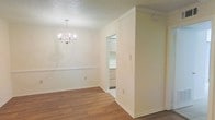This single-story space presents a clean, neutral aesthetic highlighted by warm wood-look laminate flooring that runs throughout the main living area. The dining area serves as a focal point, featuring an elegant multi-arm chandelier suspended from a textured popcorn ceiling, adding a touch of classic charm. Crown molding frames the upper walls, lending a refined architectural detail to the otherwise understated interior.

The walls are finished in a soft, creamy white tone, complemented by white baseboards that create a cohesive and tidy appearance. A chair rail molding detail runs along one wall of the dining area, adding subtle visual interest and a traditional design element.

An open doorway visible from the main area leads to an adjacent room — likely a kitchen or utility space — where built-in cabinetry can be glimpsed, suggesting practical storage solutions. The transition between spaces is defined by a structural wall partition, creating distinct zones while maintaining an open sense of flow.

Overall, the space offers a bright, well-maintained environment with classic detailing, neutral finishes, and a functional layout well-suited for everyday living.