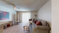 Located on Atlantic Avenue in Long Beach, California, this single-floor unit offers a practical and efficiently designed layout. Light wood laminate flooring runs throughout, creating a cohesive and clean aesthetic across all areas of the space.

The living room serves as the heart of the unit, featuring a textured ceiling, a ceiling fan with integrated lighting, and a large sliding glass door dressed with vertical blinds that allows natural light to fill the room. The open layout accommodates comfortable seating and provides enough room for decorative accents, lending a relaxed, casual ambiance.

The kitchen is positioned adjacent to the living area in an open-concept arrangement. It is equipped with white cabinetry, a tile countertop with a peninsula overhang, a white range, and a full-size refrigerator. The consistent white finish throughout the kitchen gives it a clean, functional appearance.

The bathroom is accessible via a short hallway and features a wide vanity with a large mirror framed by overhead lighting, white cabinetry with drawer storage, and the same light wood flooring found throughout the unit. A closet with mirrored sliding doors is located just outside the bathroom, adding practical storage to the space.

Overall, the unit presents a straightforward, well-maintained interior with a neutral color palette that creates a bright and inviting atmosphere throughout.