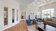 Located at 8234 Omaha Cir in Spring Hill, Florida, this single-story community clubhouse is a welcoming and thoughtfully designed gathering space managed by HTG Management. The interior is defined by dramatically high ceilings and an open floor plan that creates a bright, airy atmosphere throughout. Warm wood-look flooring runs continuously across the space, complementing the clean, neutral color palette of white and soft gray walls.

The main lounge area serves as the heart of the clubhouse, furnished with plush blue accent chairs, contemporary gray sofas, and sleek side tables arranged around a central coffee table — ideal for relaxed socializing or casual meetings. Recessed lighting and linear ceiling fixtures provide well-distributed illumination, enhancing the modern aesthetic.

A dining area features a round wood-top table surrounded by sculptural gray chairs, positioned beneath floor-to-ceiling windows that frame lush green lawns, palm trees, and a fenced waterfront view. This abundance of natural light is a defining characteristic of the space, with oversized windows lining multiple walls throughout.

Additional amenities include a private office or meeting room accessible through glass-panel French doors, and a conveniently located dual drinking fountain alcove. Large glass doors open directly to an outdoor terrace, seamlessly connecting the interior to the scenic surroundings.