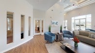 Located at 8234 Omaha Cir in Spring Hill, Florida, this single-story community clubhouse is a welcoming and thoughtfully designed gathering space managed by HTG Management. The interior is defined by dramatically high ceilings and an open floor plan that creates a bright, airy atmosphere throughout. Warm wood-look flooring runs continuously across the space, complementing the clean, neutral color palette of white and soft gray walls.

The main lounge area serves as the heart of the clubhouse, furnished with plush blue accent chairs, contemporary gray sofas, and sleek side tables arranged around a central coffee table — ideal for relaxed socializing or casual meetings. Recessed lighting and linear ceiling fixtures provide well-distributed illumination, enhancing the modern aesthetic.

A dining area features a round wood-top table surrounded by sculptural gray chairs, positioned beneath floor-to-ceiling windows that frame lush green lawns, palm trees, and a fenced waterfront view. This abundance of natural light is a defining characteristic of the space, with oversized windows lining multiple walls throughout.

Additional amenities include a private office or meeting room accessible through glass-panel French doors, and a conveniently located dual drinking fountain alcove. Large glass doors open directly to an outdoor terrace, seamlessly connecting the interior to the scenic surroundings.