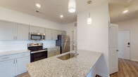 Located at 115 Beaumont Oaks Dr in Wilmington, NC, this single-floor residence offers a thoughtfully designed open-concept layout with modern finishes throughout. The kitchen serves as a central highlight, featuring granite countertops, a large peninsula island with an undermount sink and pendant lighting, gray shaker-style cabinetry, a white subway tile backsplash, and a full suite of stainless steel appliances including a range, microwave, and refrigerator. Durable wood-look plank flooring extends throughout the main living areas, complementing the clean, neutral color palette. The open living and dining area is bathed in natural light from a glass door that leads to the exterior, and is accented by a ceiling fan and a chandelier overhead. The bedroom features plush wall-to-wall carpeting and ample space. The bathroom is finished with a granite-topped vanity, gray shaker cabinetry, brushed nickel fixtures, and a full mirror. A dedicated laundry room houses a full-size top-loading washer and dryer with overhead wire shelving for additional storage. The overall ambiance is clean, bright, and move-in ready, reflecting a contemporary residential aesthetic well-suited to comfortable everyday living.