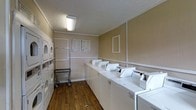 The Landry Facilities offer a practical, well-organized shared laundry space designed to accommodate multiple users efficiently. The single-floor room features a row of top-load washing machines mounted on a continuous white countertop along one wall, providing a convenient surface for sorting and folding. Stacked dryer units are positioned along the opposite wall, maximizing the use of vertical space and increasing drying capacity without expanding the room's footprint. A rolling laundry cart is available for transporting loads with ease. The room is finished with warm-toned walls and durable wood-look flooring that is easy to maintain. Overhead fluorescent lighting ensures the space is well-lit throughout. A textured ceiling adds a subtle detail to the utilitarian environment. Informational notices are posted near the entrance, providing users with guidelines and instructions for proper use of the equipment. The layout is thoughtfully arranged to allow comfortable movement between machines, making the facility functional and user-friendly for all residents who rely on it for their daily laundry needs.