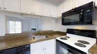 This single-floor residence offers a thoughtfully designed layout that makes efficient use of its space. The kitchen serves as a standout feature, showcasing rich granite-style countertops that contrast elegantly with crisp white cabinetry. Upper and lower cabinets provide generous storage, while a stainless steel sink with a gold-toned faucet adds a refined touch. The kitchen is equipped with a full suite of black appliances, including a Whirlpool over-the-range microwave, an electric coil range, and a dishwasher, creating a cohesive and functional cooking environment. A pass-through window connects the kitchen to an adjacent living area, enhancing the open feel of the home and allowing natural sightlines into the living space, where a fireplace is visible in the background. The white walls and clean finishes throughout contribute to a bright, airy atmosphere. The overall design strikes a balance between practicality and comfort, with classic details and durable materials that make this a welcoming and easy-to-maintain home.