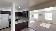 This single-story unit offers two bedrooms and one bathroom within a practical, well-designed floor plan. The kitchen features dark espresso cabinetry that extends to the upper walls, providing generous storage capacity. A stone-toned tile backsplash complements the granite-style countertops, while a stainless steel sink is positioned along the peninsula, which serves as a natural divider between the kitchen and the adjacent living area. The flooring in the kitchen is a wood-style hard surface, offering easy maintenance.

The living area transitions seamlessly from the kitchen and is finished with wall-to-wall carpeting, lending a warm, comfortable feel to the space. A window along the far wall allows natural light to filter in, brightening the room throughout the day. A wall-mounted air conditioning unit provides climate control. An electrical panel is also accessible within the living area. Recessed lighting in the kitchen and ceiling-mounted fixtures in the living room ensure adequate illumination throughout. The neutral wall tones throughout create a clean, versatile backdrop suitable for a variety of interior styles.