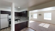 This single-story unit offers two bedrooms and one bathroom within a practical, well-designed floor plan. The kitchen features dark espresso cabinetry that extends to the upper walls, providing generous storage capacity. A stone-toned tile backsplash complements the granite-style countertops, while a stainless steel sink is positioned along the peninsula, which serves as a natural divider between the kitchen and the adjacent living area. The flooring in the kitchen is a wood-style hard surface, offering easy maintenance.

The living area transitions seamlessly from the kitchen and is finished with wall-to-wall carpeting, lending a warm, comfortable feel to the space. A window along the far wall allows natural light to filter in, brightening the room throughout the day. A wall-mounted air conditioning unit provides climate control. An electrical panel is also accessible within the living area. Recessed lighting in the kitchen and ceiling-mounted fixtures in the living room ensure adequate illumination throughout. The neutral wall tones throughout create a clean, versatile backdrop suitable for a variety of interior styles.