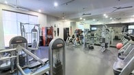 This fitness center is a fully equipped, single-floor gym designed to support a comprehensive workout experience. The space features a durable dark rubberized floor throughout, providing a safe and practical surface for a wide variety of exercises. A broad selection of strength training equipment and resistance machines lines the space, complemented by cardio equipment including stationary bikes and other Star Trac branded machines. A dedicated free weights area with a dumbbell rack rounds out the strength training offerings. Large wall-mounted mirrors along one side of the gym visually expand the space and allow users to monitor their form during workouts. The ceiling is fitted with recessed lighting and track lighting, ensuring the space is well-lit at all times, while ceiling fans help maintain comfortable air circulation throughout. A mounted flat-screen television provides entertainment for those engaged in longer workout sessions. For added convenience, a Coca-Cola vending machine is available on-site. Natural light filters in through windows along one wall, contributing to an open and energizing atmosphere. The overall design is clean, modern, and functional, making it a practical and well-appointed facility for fitness enthusiasts.