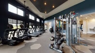 Located at 9295 One Deerfield Place in Mason, Ohio, this single-floor fitness and amenity facility offers a thoughtfully designed space for both exercise and relaxation. The expansive fitness area is defined by deep teal accent walls, soaring ceilings, and geometric-patterned carpet tiles that add a contemporary aesthetic throughout. A generous row of True-brand treadmills and ellipticals lines the perimeter, facing large windows that frame outdoor views and fill the space with natural light. A dedicated strength training zone features cable machines, multi-use weight stations, free weights, medicine balls, and resistance bands, complemented by full-length mirrors that enhance the sense of openness. Ceiling fans and recessed lighting maintain a comfortable, well-lit environment. Adjacent to the fitness floor, a refined lounge area provides a welcoming retreat. This space showcases an ornate coffered ceiling with a sculptural honeycomb design, a wood-paneled accent wall housing a flat-screen television, and built-in shelving with a beverage cooler. Plush leather armchairs surround a round coffee table atop a patterned area rug, while black-framed glass partitions create an elegant visual boundary. A water fountain station serves both areas, completing this well-appointed amenity facility.