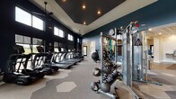 Located at 9295 One Deerfield Place in Mason, Ohio, this single-floor fitness and amenity facility offers a thoughtfully designed space for both exercise and relaxation. The expansive fitness area is defined by deep teal accent walls, soaring ceilings, and geometric-patterned carpet tiles that add a contemporary aesthetic throughout. A generous row of True-brand treadmills and ellipticals lines the perimeter, facing large windows that frame outdoor views and fill the space with natural light. A dedicated strength training zone features cable machines, multi-use weight stations, free weights, medicine balls, and resistance bands, complemented by full-length mirrors that enhance the sense of openness. Ceiling fans and recessed lighting maintain a comfortable, well-lit environment. Adjacent to the fitness floor, a refined lounge area provides a welcoming retreat. This space showcases an ornate coffered ceiling with a sculptural honeycomb design, a wood-paneled accent wall housing a flat-screen television, and built-in shelving with a beverage cooler. Plush leather armchairs surround a round coffee table atop a patterned area rug, while black-framed glass partitions create an elegant visual boundary. A water fountain station serves both areas, completing this well-appointed amenity facility.
