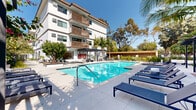 Located at 8501 S Sepulveda Blvd in Los Angeles, this thoughtfully designed common area offers a seamless blend of indoor comfort and outdoor California living. The interior living room features light-toned tile flooring, white painted brick accent walls, and recessed lighting that keeps the space bright and airy. A large sliding glass door connects the interior directly to the outdoor pool area, flooding the room with natural light and providing unobstructed views of the resort-style amenities. The living room is furnished with contemporary pieces, including a plush blue sofa and lounge chair, a sleek media console, and carefully curated décor that strikes a balance between modern and welcoming. A built-in kitchenette area showcases light wood cabinetry, a marble-style backsplash, and open shelving, further enhancing the functionality of the space. Outside, a generously sized swimming pool is surrounded by a wide concrete deck lined with navy blue chaise lounges, a shaded pergola seating area, and lush tropical landscaping including towering palm trees. The multi-story white stucco building with warm wood balcony accents frames the courtyard beautifully, creating a cohesive and resort-inspired outdoor retreat under the sunny Los Angeles sky.