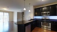 This single-floor space showcases a thoughtfully designed open-concept layout that seamlessly blends style and functionality. The kitchen serves as the centerpiece of the home, featuring rich dark cabinetry that contrasts beautifully against light walls, creating a bold yet sophisticated aesthetic. Dark granite countertops extend across ample workspace, continuing onto a curved kitchen island that provides additional prep space and casual seating potential. Two elegant bell-shaped pendant lights hang overhead, casting warm, focused illumination over the island. Stainless steel appliances — including a gas range, built-in microwave, and dishwasher — lend a professional, polished feel to the culinary space. A stainless steel undermount sink is integrated into the L-shaped counter, completing the kitchen's practical design. Warm hardwood flooring flows throughout, adding natural warmth that softens the kitchen's darker tones. The open layout connects the kitchen to adjacent living areas, with glimpses of a hallway, doors, and a utility area visible in the background, suggesting a well-organized and efficiently planned floor plan. The overall ambiance is contemporary and clean, offering a comfortable and visually cohesive living environment.