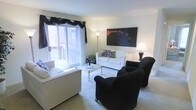 This single-floor space offers a welcoming and well-appointed interior designed for comfortable everyday living. The living room serves as the heart of the space, bathed in natural light that streams through a large sliding glass door dressed with dramatic black drape accents. The glass door opens directly onto a private balcony, providing an easy indoor-outdoor connection and pleasant outdoor views. Soft, neutral carpeting runs throughout the living area, adding warmth underfoot and contributing to the room's relaxed ambiance. The seating arrangement is thoughtfully composed, featuring a white sofa accented with patterned throw pillows, complemented by a pair of bold black armchairs that create a striking visual contrast. A sleek glass-top coffee table anchors the seating area, while a tall floor lamp and a classic table lamp provide layered ambient lighting for the evenings. A large abstract canvas painting in black and red tones hangs prominently on the main wall, adding an artistic focal point that ties the room's color palette together. The overall aesthetic blends contemporary style with inviting comfort, making this a versatile and functional living space.