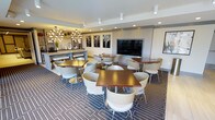 This single-floor communal space is thoughtfully designed to serve as a social hub, combining the functionality of a dining area and lounge. The room features several rich, dark-topped tables paired with contemporary cream-colored chairs arranged throughout, accommodating multiple groups simultaneously. A patterned carpet in shades of navy and grey anchors the seating area, adding visual interest underfoot.

A prominent large-screen display is built into a sleek, light-toned wall unit, making the space well-suited for presentations or entertainment. Overhead, a combination of recessed lighting and a striking sputnik-style chandelier provides layered illumination, contributing to both functionality and ambiance.

Along one side of the room, a bar or serving counter stretches beneath open shelving, accented with decorative objects and ambient lighting. The counter's light finish contrasts nicely with the darker furnishings throughout the space. Framed wall art featuring tree branch motifs adds a refined, nature-inspired decorative touch. The overall aesthetic is modern and polished, creating a welcoming environment suitable for dining, socializing, or community events.