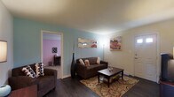 This single-floor space centers around a welcoming living area distinguished by rich, dark hardwood flooring and soft blue-painted walls that create a calm, relaxed atmosphere. The living room is furnished with a dark brown sofa accented by decorative throw pillows, paired with a matching coffee table atop a patterned area rug that adds warmth and visual interest to the space. A tall decorative vase with slender branches serves as an elegant floor accent, while wall-mounted mirrored wave décor and a colorful vintage-style painting add personality to the room. A floor lamp provides warm ambient lighting near the seating area. White trim and baseboards offer a clean contrast against the colored walls and dark flooring throughout. An open doorway leads to an adjacent room featuring distinctly lavender-pink walls and carpeted flooring, suggesting a bedroom or secondary living space. A small console table and additional furnishings are visible within that room. The entry door, positioned to the right of the living area, is framed with white molding, reinforcing the tidy, well-maintained character of this single-level space.