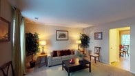 This two-story residence offers a thoughtfully arranged layout across two levels, combining comfort and classic style. The living room serves as a welcoming centerpiece, featuring light-toned walls that reflect warm ambient lighting from elegant table lamps positioned on either side of the space. A plush, neutral-toned sofa adorned with rich burgundy and patterned accent pillows anchors the seating area, complemented by a dark wood coffee table that adds contrast to the light hardwood flooring beneath. Framed artwork decorates the walls, adding a curated, homey touch to the room. Lush indoor potted trees flank the space, introducing a refreshing natural element that softens the interior. A cushioned side chair and matching end tables complete the living area's furnishings. The textured ceiling adds subtle architectural character, while the open, airy feel of the room contributes to an overall sense of spaciousness. Crown molding along the upper walls provides a clean, finished look throughout. The home's two-floor configuration offers distinct zones for living and private spaces, making it a functional and comfortable residence suited for everyday living.