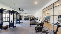 This modern fitness center offers a comprehensive workout environment thoughtfully designed to accommodate a wide range of exercise disciplines. The open-plan layout provides ample floor space for both cardio and strength training, finished with durable, multi-toned carpet tile throughout. Along one side, a row of treadmills and stationary bikes — including smart-screen-equipped models — lines the wall, accompanied by an elliptical and air bike for varied cardio options. The strength training area features a full dumbbell rack, adjustable benches, a multi-functional cable training rig, and a dedicated weight machine section, all reflected in a large wall-mounted mirror that enhances the sense of space. A standout architectural feature is the roll-up glass garage door that opens directly to an outdoor terrace area, flooding the space with natural light and enabling indoor-outdoor workouts. Ceiling fans circulate air throughout the facility, contributing to a comfortable training environment. A separate glass-partitioned room, framed in black steel, provides an additional workout or stretching area. The space is punctuated by a bold motivational mural reading "TRAIN HARD," adding an energetic, design-forward element to the overall ambiance. A water fountain is also conveniently accessible within the space.