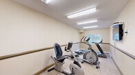 Located at 30 Nevada Pl in St. Albert, Alberta, this dedicated exercise room provides a practical and well-equipped fitness space. The room features light-toned walls with a continuous wooden handrail running along the perimeter — a thoughtful accessibility feature that adds both safety and a warm, structured aesthetic. Flooring throughout is covered in neutral carpet tiles, offering comfort underfoot during workouts.

The room is furnished with a variety of cardio equipment, including a treadmill, an elliptical trainer, a recumbent stationary bike, and an upright stationary bike, along with a mini trampoline and exercise balls for supplementary training. A wall-mounted flat-screen television is positioned for easy viewing during exercise sessions.

Overhead, flush fluorescent lighting panels provide bright, even illumination throughout the space. A window fitted with horizontal blinds allows natural light to enter while maintaining privacy. A hand sanitizer dispenser mounted near the entrance reflects attention to hygiene. The textured ceiling complements the room's clean, functional design. Overall, this exercise room is efficiently organized to offer a comprehensive range of cardio fitness options within a compact, accessible, and well-lit environment.