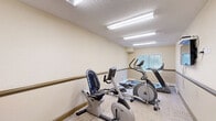 Located at 30 Nevada Pl in St. Albert, Alberta, this dedicated exercise room provides a practical and well-equipped fitness space. The room features light-toned walls with a continuous wooden handrail running along the perimeter — a thoughtful accessibility feature that adds both safety and a warm, structured aesthetic. Flooring throughout is covered in neutral carpet tiles, offering comfort underfoot during workouts.

The room is furnished with a variety of cardio equipment, including a treadmill, an elliptical trainer, a recumbent stationary bike, and an upright stationary bike, along with a mini trampoline and exercise balls for supplementary training. A wall-mounted flat-screen television is positioned for easy viewing during exercise sessions.

Overhead, flush fluorescent lighting panels provide bright, even illumination throughout the space. A window fitted with horizontal blinds allows natural light to enter while maintaining privacy. A hand sanitizer dispenser mounted near the entrance reflects attention to hygiene. The textured ceiling complements the room's clean, functional design. Overall, this exercise room is efficiently organized to offer a comprehensive range of cardio fitness options within a compact, accessible, and well-lit environment.