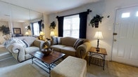 Located at 454 Heather Dr in Mantua, New Jersey, this single-floor home offers a welcoming and well-appointed living environment. The living room serves as the heart of the space, showcasing a warm, neutral color palette with light-toned walls and plush carpeting underfoot. A comfortable upholstered sofa and coordinating ottoman are centered around a wooden coffee table with wrought-iron accents, creating an ideal gathering area. Ambient lighting is provided by classic table lamps that cast a soft, inviting glow throughout the room. A large mirror on one wall enhances the sense of openness, reflecting light and adding depth to the interior. Windows dressed with dark curtain panels allow for natural light during the day while offering privacy in the evenings. Decorative touches such as hanging potted plants and carefully selected accessories add character and a lived-in charm to the space. The overall ambiance is warm, cozy, and thoughtfully arranged, making this single-level home a comfortable retreat suited for everyday living in the quiet residential community of Mantua, NJ.