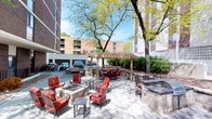 This inviting outdoor space is designed for relaxation and social gatherings, offering a thoughtfully landscaped patio environment within an urban setting. The space is anchored by two prominent stone-built fire pits, each surrounded by cushioned wrought-iron chairs with bold red upholstery, creating warm and welcoming conversation areas. A wooden pergola serves as a central architectural focal point, providing partial shade over a dining area furnished with a table and matching red-cushioned seating. The patio surface is finished with neatly laid pavers, adding texture and a polished aesthetic throughout. Raised stone planter walls define the perimeter of the space, offering a sense of enclosure while maintaining open sightlines to the surrounding greenery. Mature deciduous trees, in full seasonal foliage, provide natural shade and an organic, park-like atmosphere that softens the urban backdrop. Additional seating areas are distributed throughout, offering flexible arrangements for both intimate gatherings and larger groups. The overall ambiance is one of casual elegance, blending durable outdoor materials with comfortable furnishings to create a functional and visually appealing communal outdoor living area.
