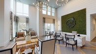 This single-floor amenity facility presents a thoughtfully designed community space that blends upscale aesthetics with practical functionality. The grand lobby immediately impresses with soaring double-height ceilings, an eye-catching cascading chandelier, and shiplap accent walls. A dramatic living moss wall art installation serves as a striking focal point near the reception desk, which features a distinctive marble-look surface. Floor-to-ceiling windows flood the space with natural light while offering outdoor views, and elegant patterned drapery adds a refined finishing touch. A comfortable lounge area with oversized armchairs and tranquil waterfront views provides an inviting retreat.

The community kitchen is a standout feature, anchored by a generous white quartz island with seating, dark navy cabinetry, a patterned tile backsplash, open wood shelving, and exposed wooden ceiling beams that add warmth and character. Large-format tile flooring grounds the space elegantly.

An airy dining room with soaring ceilings, bold circular pendant lighting, and black-framed French doors opening to an outdoor courtyard accommodates group gatherings with ease. The fitness center is well-equipped with cardio machines, free weights, exercise balls, and a full-length mirror wall, all bathed in natural light through frosted windows. Black-framed glass doors connect the amenity areas cohesively throughout.