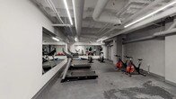 Located at 8121 Van Nuys Blvd in the Panorama City neighborhood of Los Angeles, this fitness center offers a well-equipped, single-floor workout space designed for functional training and cardio exercise. The gym features an open-plan layout with a spacious floor area finished in durable rubber flooring, ideal for high-traffic athletic use. The industrial-style exposed ceiling showcases a network of white-painted ductwork and piping, complemented by linear LED strip lighting that ensures bright, even illumination throughout the space. Large wall-mounted mirrors span multiple sections of the gym, creating a sense of expanded space and allowing users to monitor their form during workouts. The cardio zone is equipped with multiple treadmills and elliptical machines, while a dedicated cycling area houses spin bikes suited for high-intensity interval training. Red exercise balls are stored along the walls, adding a pop of color to the otherwise neutral gray-and-white palette. Structural concrete columns punctuate the floor plan, and the walls feature a smooth gray finish that contributes to the facility's clean, modern aesthetic. The ceiling-mounted ventilation system supports air circulation across the entire space. Overall, this fitness center provides a comprehensive and efficiently organized environment for a variety of workout routines.