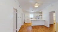 This single-floor space features an open-plan layout that seamlessly connects the living area to the kitchen, creating a spacious and airy feel. The kitchen is outfitted with crisp white cabinetry, white appliances including a refrigerator, electric range, and dishwasher, as well as a double sink positioned beneath a window that allows natural light to fill the space. A peninsular breakfast bar extends from the kitchen, offering additional counter space and a casual dining option. The flooring throughout is warm-toned hardwood-style, adding a welcoming contrast to the all-white walls, cabinetry, and textured ceiling. Vaulted ceiling lines lend an elevated sense of height to the open area, while a ceiling fan with an integrated light fixture and a separate track lighting system provide both ambient and task lighting. Architectural details such as raised ceiling transitions between areas give the space subtle definition. A hallway leads to additional rooms, with a louvered closet door visible, suggesting convenient storage. Overall, the space presents a clean, bright, and functional aesthetic.