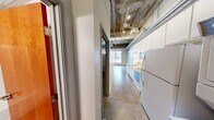 This single-floor urban loft unit showcases a distinctive industrial aesthetic that blends raw architectural character with functional living. Throughout the space, exposed concrete ceilings with visible ductwork and track lighting create an open, warehouse-inspired atmosphere, while polished concrete floors tie the rooms together cohesively.

The main living area is bright and open, benefiting from large multi-pane windows that invite generous natural light and offer views of the surrounding urban streetscape. A wall-mounted mini-split system provides efficient climate control. The open layout allows for flexible furniture arrangements.

The kitchen runs along one wall in a galley-style configuration, featuring white cabinetry, granite countertops, and a bold yellow subway tile backsplash that adds a vibrant pop of color. Appliances include an electric range, over-the-range microwave, refrigerator, and dishwasher. The kitchen opens directly toward the main living space, reinforcing the unit's open feel.

The bathroom is accessed through a warm-toned solid wood door, offering a clean and functional design with neutral gray tile flooring, a full tub-and-shower combination, and white fixtures. The space is straightforward and well-appointed.

Overall, this unit delivers an urban loft character defined by its raw industrial finishes, natural light, and efficient use of space.