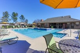 This outdoor amenity area in Phenix City, Alabama, centers around a generously sized swimming pool with clear, blue water and a connected spa or wading feature along one side. The pool deck is finished in light-colored concrete and offers ample space for lounging, furnished with multiple reclining chairs and umbrella-shaded seating areas, creating a well-appointed environment for outdoor relaxation. A wrought-iron perimeter fence encloses the pool area, providing both security and a view of the surrounding landscaping, including mature trees and maintained shrubbery. The adjacent single-story brick building, featuring a pitched shingle roof, large windows, and glass doors, serves as a clubhouse or amenity facility. This structure opens directly onto the pool deck, facilitating convenient access between indoor and outdoor spaces. The overall setting is bright and open, benefiting from abundant natural light under clear skies. Brick residential buildings are visible in the background, contributing to the cohesive architectural character of the community. The combination of functional amenities, durable materials, and thoughtful layout makes this outdoor space a notable communal feature of the Phenix City property.