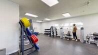 Located at 607 Palm Glades Dr in Belle Glade, Florida, this fitness facility offers a spacious, single-floor layout designed to accommodate a full range of workout needs. The interior features crisp white walls and durable rubber-speckled flooring throughout, creating a clean and functional environment suited for both casual and dedicated training.

The cardio section is well-appointed with multiple treadmills, an elliptical trainer, and stationary bikes, each positioned beneath wall-mounted flat-screen televisions for an engaging workout experience. Natural light filters in through several windows along the perimeter walls, offering views of the surrounding Florida landscape and contributing to an open, airy atmosphere.

The strength training area is equally comprehensive, featuring a multi-station cable machine, adjustable benches, and an extensive selection of dumbbells arranged on tiered racks. A full-length mirror and colorful weight plates on a barbell rack add both practicality and visual interest to the space. Recessed panel lighting throughout ensures consistent, bright illumination across the entire facility.

Additional equipment such as cable pull machines and seated resistance units round out the offerings, making this a versatile, fully equipped fitness space suitable for a wide variety of training programs.