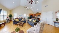 This single-floor community facility is thoughtfully designed to offer both comfort and functionality across its key areas. The heart of the space is a grand open-concept great room distinguished by soaring vaulted ceilings adorned with multiple ceiling fans and chandelier lighting. Warm hardwood flooring flows throughout, and abundant natural light pours in through numerous windows. The living area comfortably accommodates multiple seating arrangements, including plush sofas and accent chairs accented with yellow throw pillows, centered around a round coffee table. The adjoining kitchen is well-appointed with rich wood cabinetry, stone countertops, tile backsplash, a central island with bar seating, and full black appliances including a refrigerator, dishwasher, and range. A dedicated dining area with wainscoting and a large wood dining table provides a more formal gathering space. A secondary lounge area with a wall-mounted television and additional seating offers a quieter retreat. The fitness center is a standout feature, boasting its own vaulted ceilings with large windows that flood the room with natural light. It is fully equipped with treadmills, an elliptical, cable weight machines, benches, and free weights, all set on durable rubberized flooring with motivational wall graphics. A glazed entry door with sidelights and a brick exterior complete this welcoming facility.