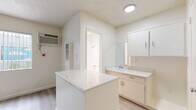Located on Magnolia Boulevard in North Hollywood, Los Angeles, this single-floor unit offers a efficiently designed layout comprising a living area and a full bathroom. The space features light gray laminate wood flooring throughout, complementing the bright, neutral-toned walls that create a clean and airy atmosphere. A textured ceiling adds subtle architectural character to the interior.

The main living area is open and versatile, with a window fitted with vertical blinds that allows natural light to filter in and offers a glimpse of greenery outside. A wall-mounted HVAC unit provides climate control. The kitchenette is defined by a white peninsula with a light stone-style countertop, paired with white upper cabinetry and a mirror-backed vanity area, maximizing both function and style in a compact footprint.

The bathroom is finished in crisp white, featuring fully tiled walls, a combination bathtub and shower with polished chrome fixtures, and a small window that introduces natural light. The overall design aesthetic is minimal and modern, with white trim and baseboards tying the space together cohesively. This unit is well-suited for those seeking a practical, move-in-ready space in the vibrant North Hollywood neighborhood of Los Angeles.